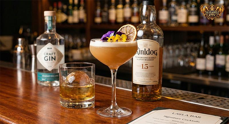 Cận cảnh một ly Thức uống pha chế signature màu vàng cam được trang trí hoa tươi và lát cam sấy trên quầy bar gỗ tại Layla Bar. Bên cạnh là ly whisky với đá tảng tròn và một chai rượu mạnh cao cấp.