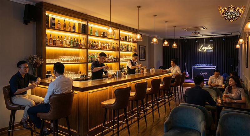 Không gian nội thất sang trọng bên trong Layla Bar. Quầy bar gỗ dài với tủ rượu lớn được chiếu sáng vàng ấm áp, bartender đang pha chế, khách hàng ngồi thưởng thức đồ uống trên ghế da cao cấp.