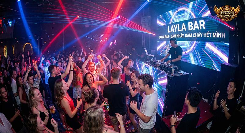 Không khí tiệc tùng đêm khuya sôi động tại Layla Bar. Đám đông giới trẻ đang nhảy múa dưới ánh đèn laser đỏ và xanh. Trên sân khấu, DJ chơi nhạc với màn hình LED lớn phía sau hiển thị dòng chữ "DÁM NHẢY, DÁM CHÁY HẾT MÌNH"