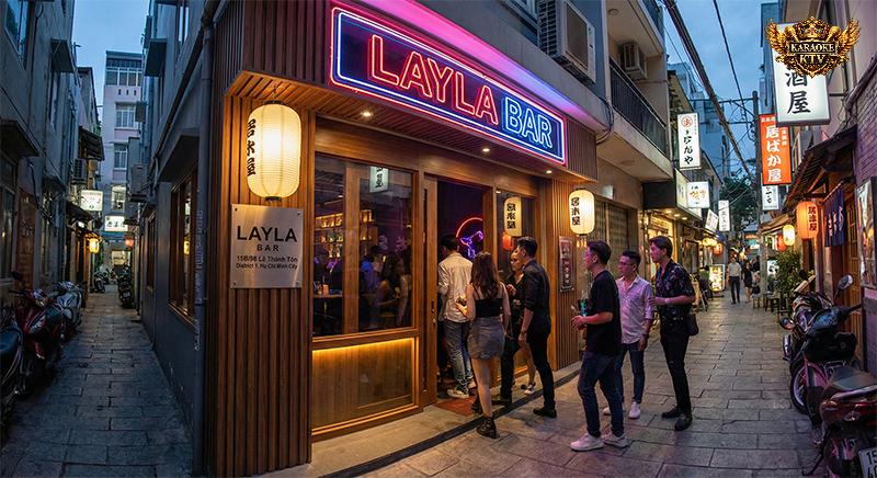 Mặt tiền Layla Bar tại hẻm 15B/98 Lê Thánh Tôn, Quận 1 vào buổi tối. Bảng hiệu neon "LAYLA BAR" sáng đèn, lồng đèn Nhật trang trí, nhóm khách trẻ đang bước vào quán.